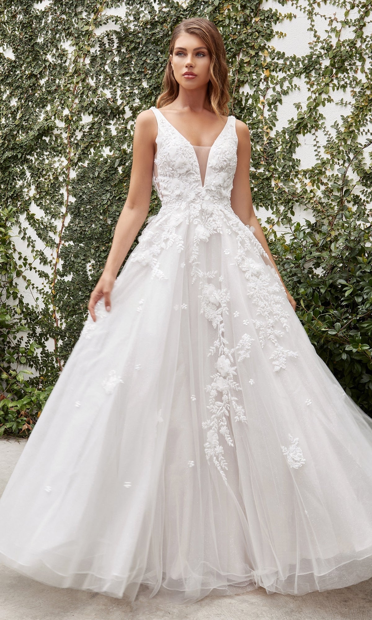 A woman wearing the French White Long Wedding Gown: Ladivine A1028W stands outdoors in front of a green ivy-covered wall.